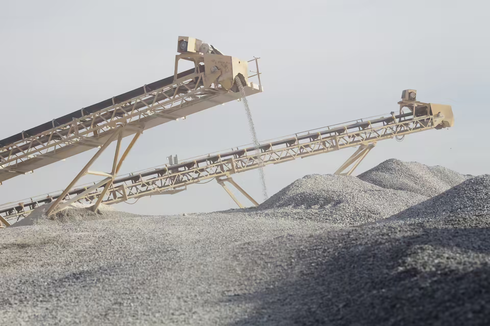 Two inclined conveyors carrying gravel over stockpiles at a quarry under a pale sky