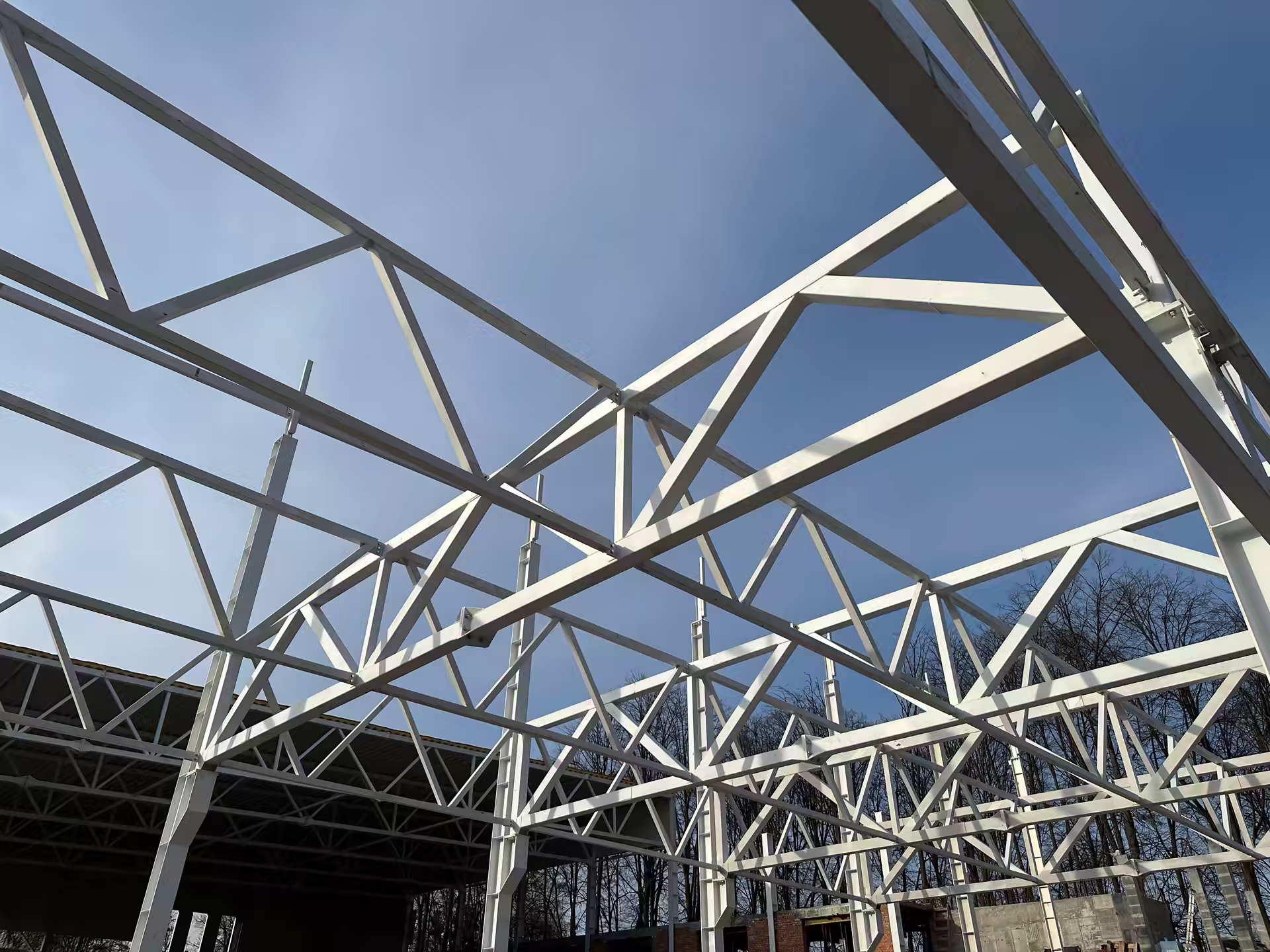 White steel roof trusses of a building under construction, photographed from below against a blue sky