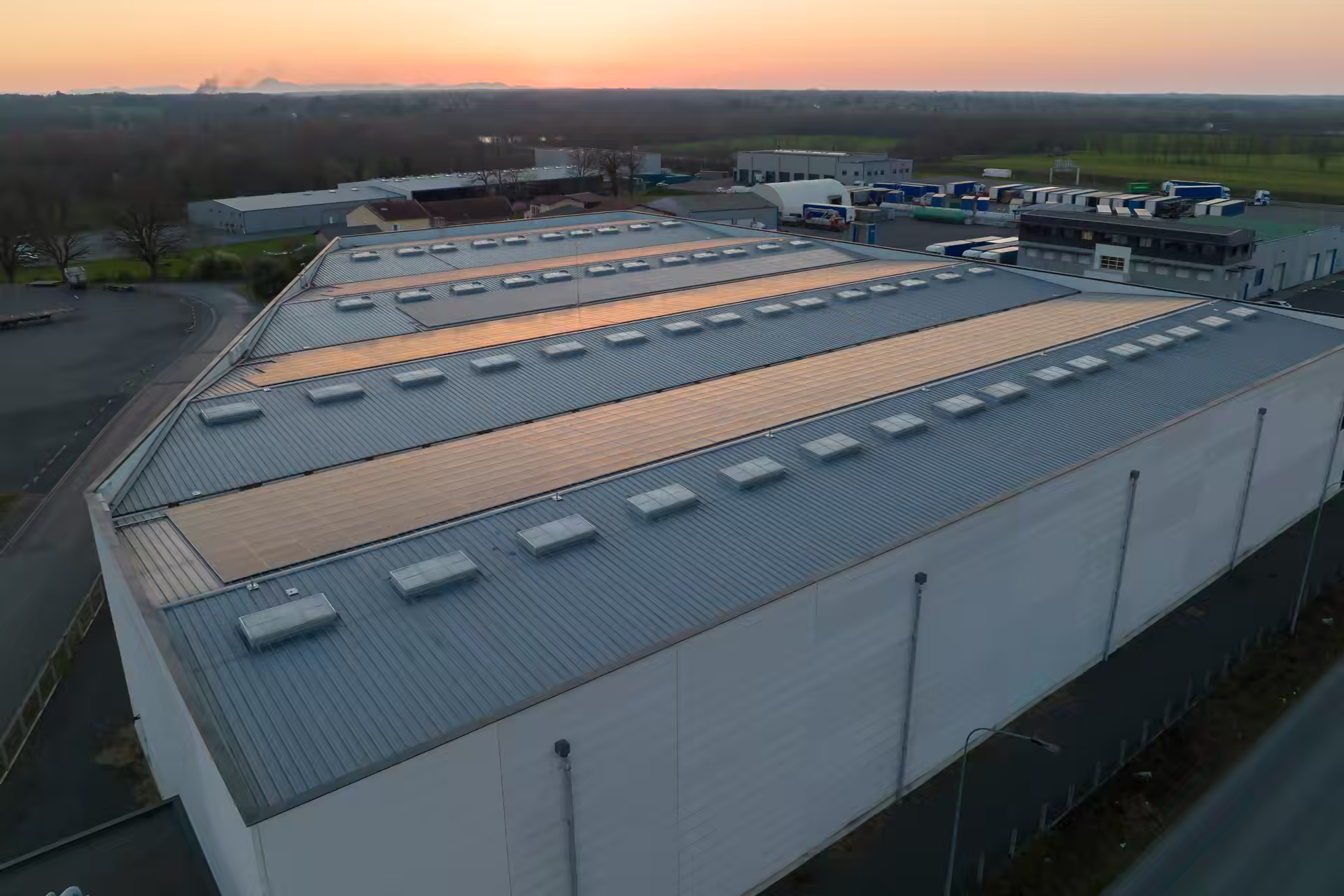 Aerial view at sunset of a long industrial building with rooftop skylights and solar panels, and a yard beyond