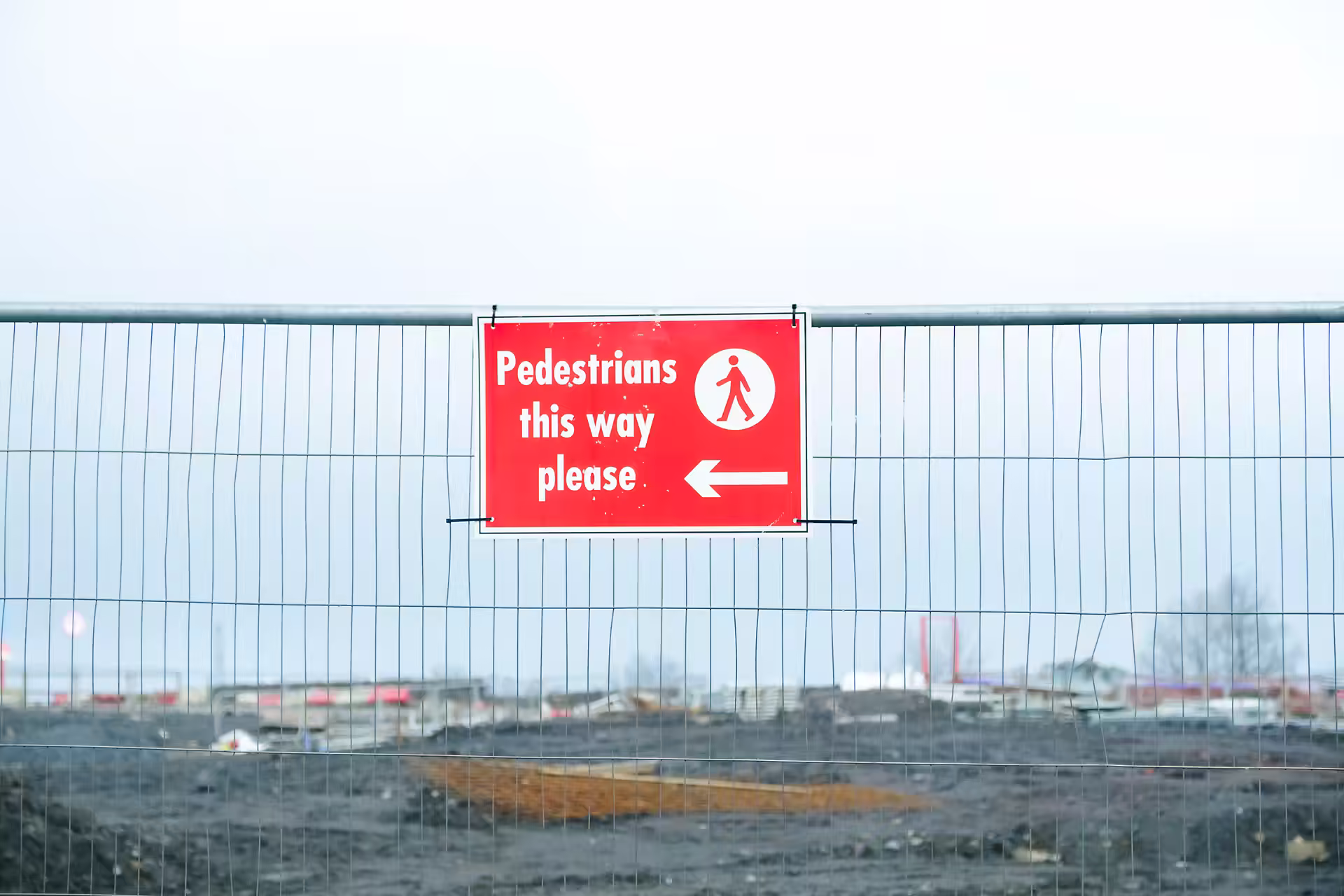 Red "Pedestrians this way please" sign on temporary fencing around a cleared construction site