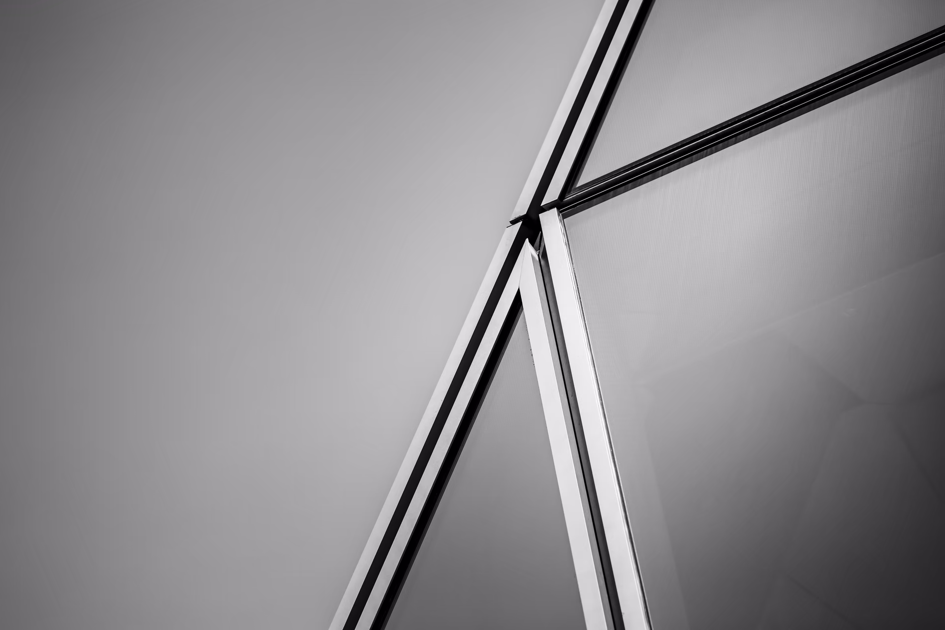 Black-and-white detail of glass facade mullions meeting at an angle against a pale grey sky
